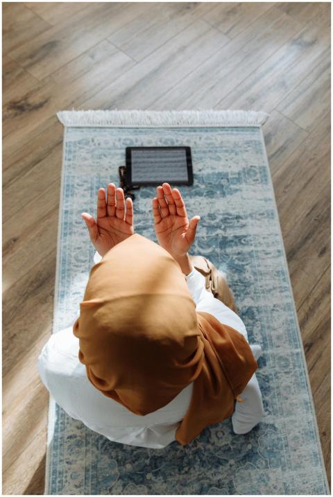 A woman in a hijab prays on a rug with a tablet ne