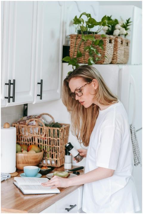 A woman multitasks in a kitchen, reading a book an
