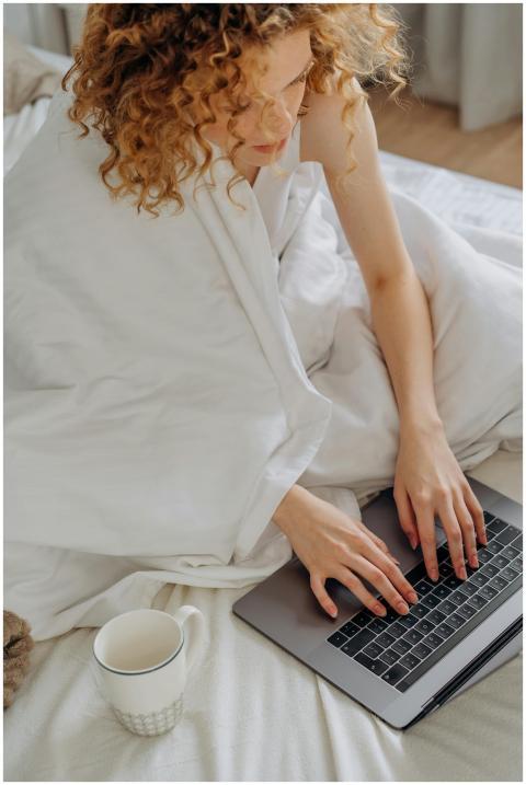 Adult woman typing on a laptop in bed with a cup o