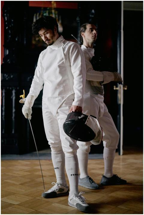 Two male fencers in white gear take a break indoor