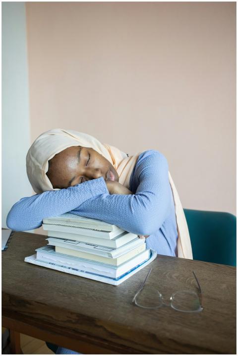 A tired woman in hijab sleeps on a stack of books,