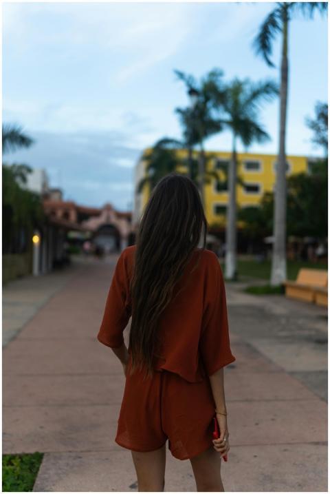 A woman with long hair walks along a palm-lined, u