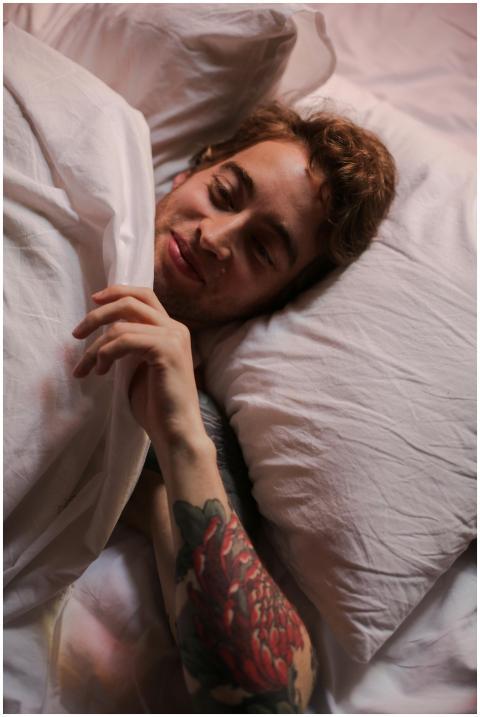 A tattooed man relaxes in bed, wrapped in white sh