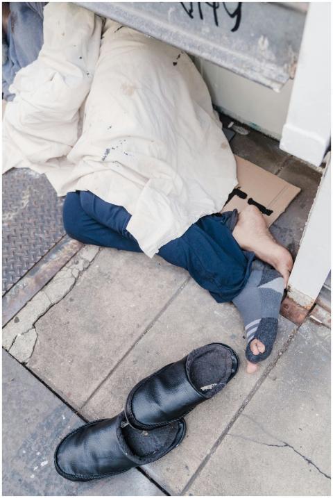 A person sleeps on the sidewalk under a blanket, h