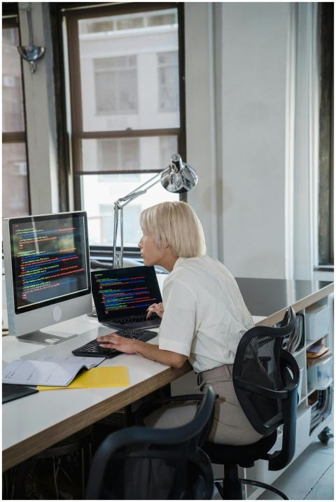 Blonde woman coding at desk with dual monitors in
