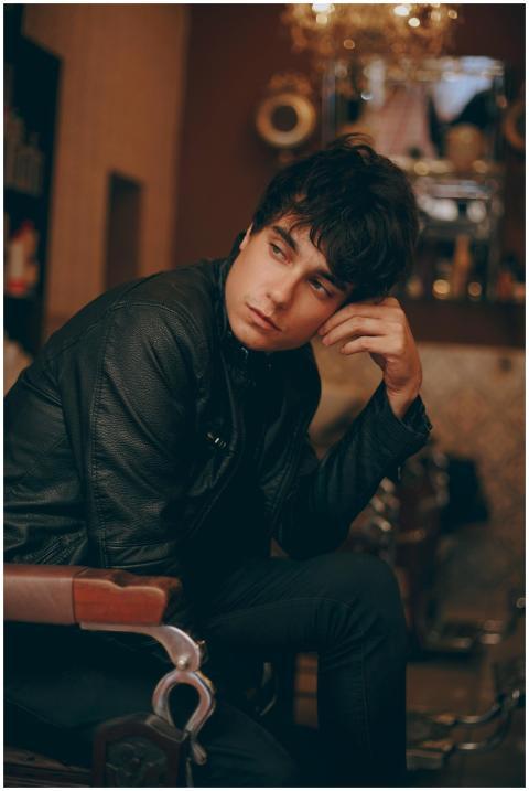 A young man seated indoors wearing a black leather