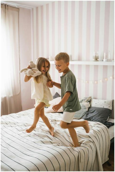 Two siblings happily jumping on a bed in a cozy ro