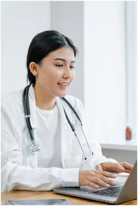 Asian female doctor in a white coat typing on a la