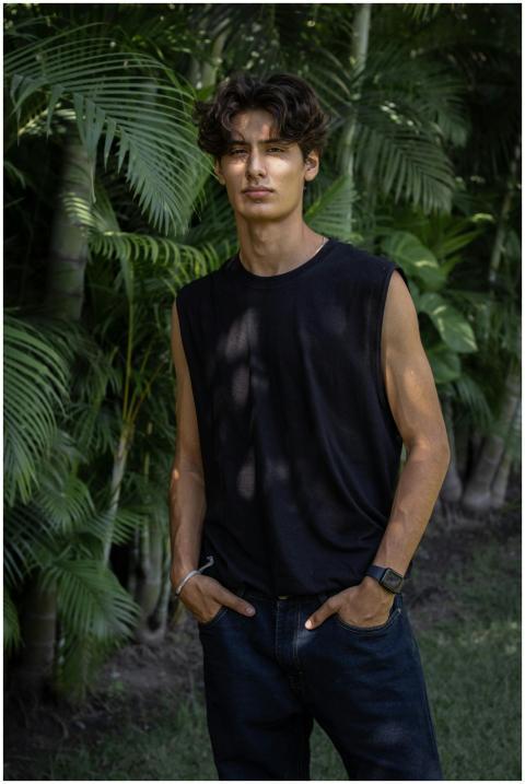 A confident young man stands outdoors with lush tr