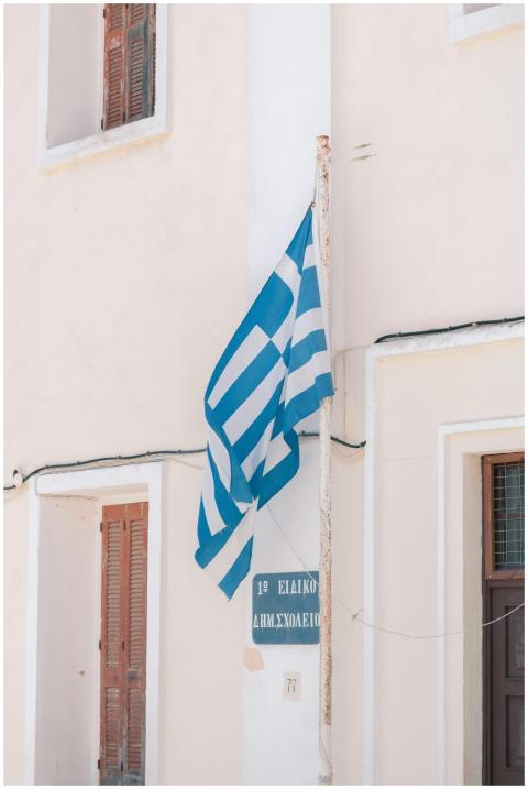 A Greek flag flutters against a white building, ca
