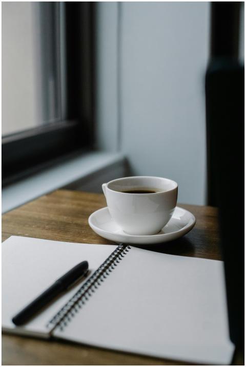 A cup of coffee and an open notebook by a window i