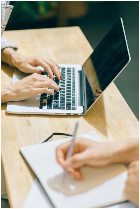 Hands typing on a laptop and writing in a notebook
