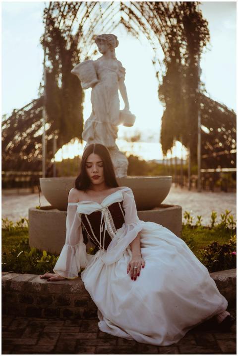 A woman in Victorian attire sits by a stone statue