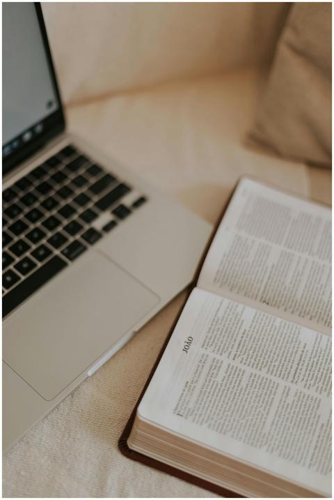 An open Bible next to a laptop on a cozy bedspread