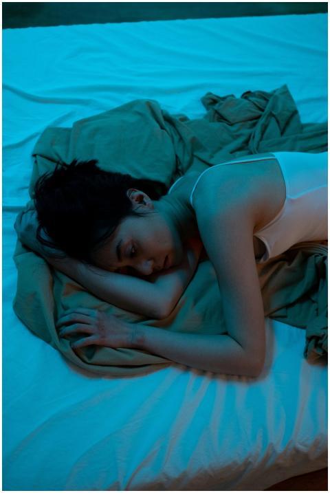 An adult woman lies on a bed in a dimly lit room,