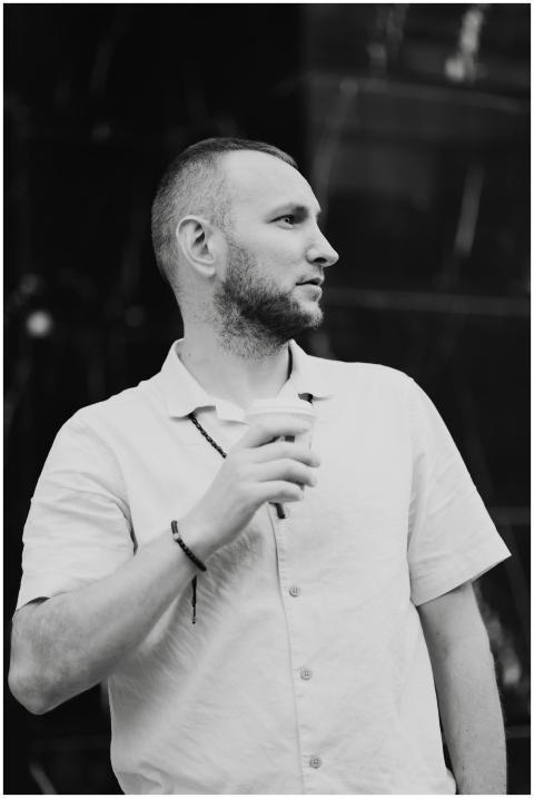 A stylish man with facial hair holds a cup, lookin