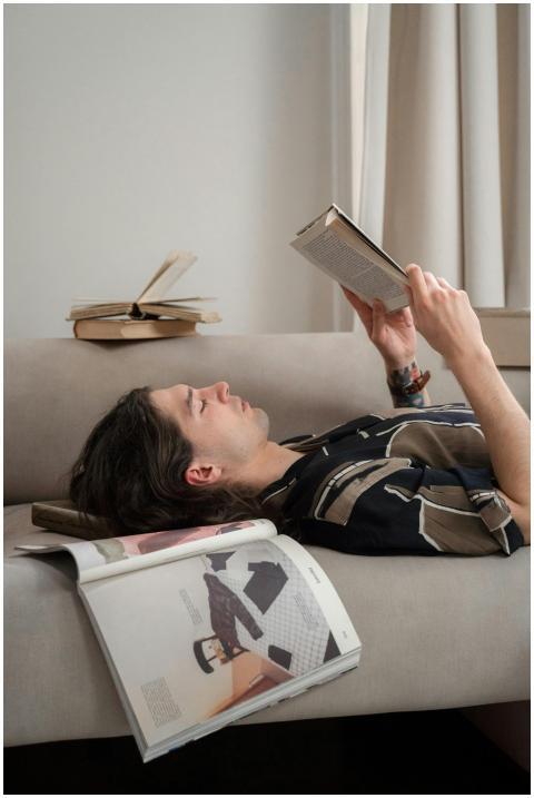 A relaxed young man lies on a sofa reading a book,