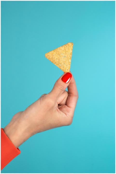 A hand with red nails holding a tortilla chip agai