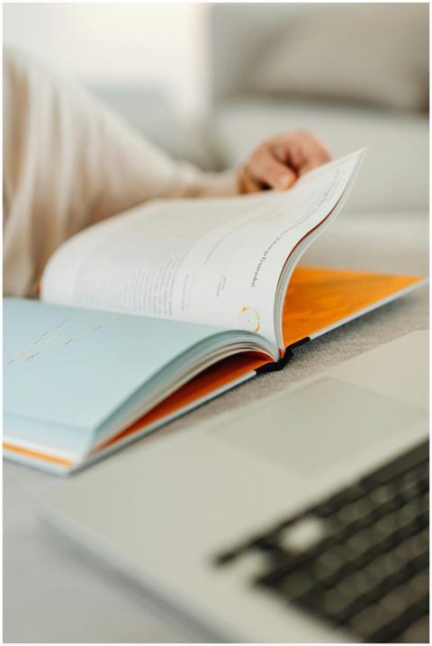 A person flips through a book beside a laptop on a