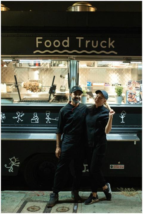 A man and woman stand in front of a food truck, of