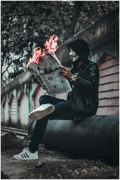 A person wearing a mask reads a flaming newspaper