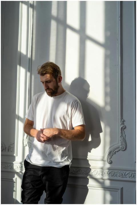 A thoughtful bearded man in a white t-shirt stands