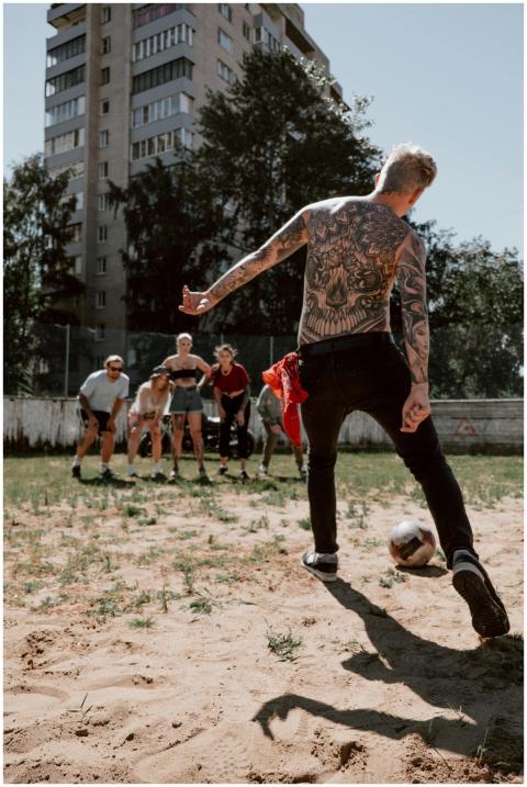 A tattooed man kicks a soccer ball on a sandlot fi