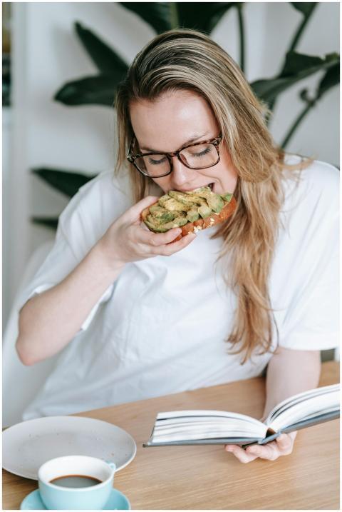 Focused female eating toast with avocado while rea