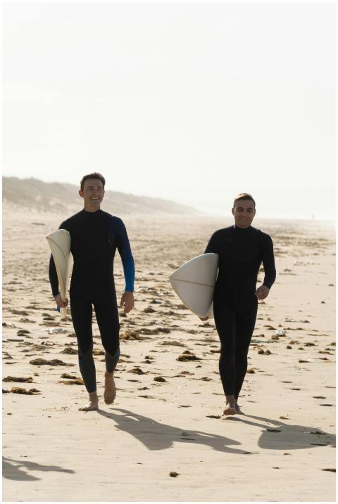 Two men in wetsuits carrying surfboards on a sunli