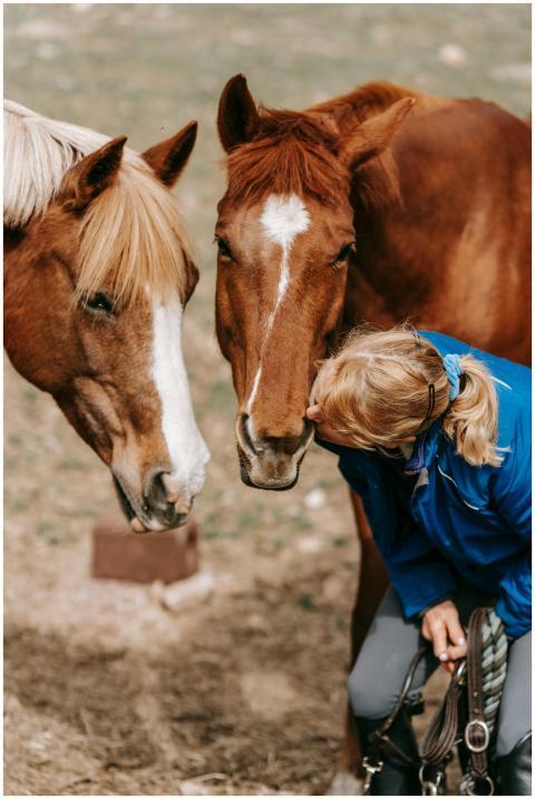 A woman lovingly kisses two brown horses outdoors,