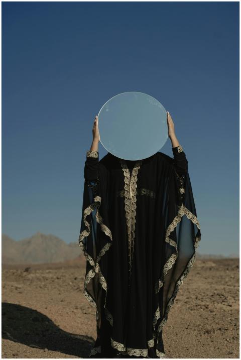 A person in a robe holds a circular mirror, reflec