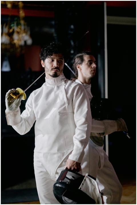 Two fencers pose aggressively with swords indoors,