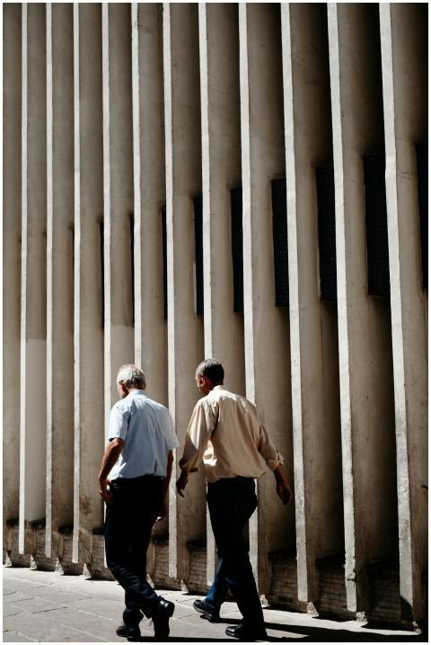 Two men walking beside a textured urban wall on a