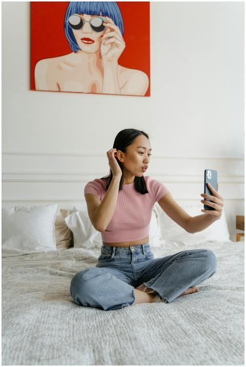 A young woman sitting on a bed taking a selfie in