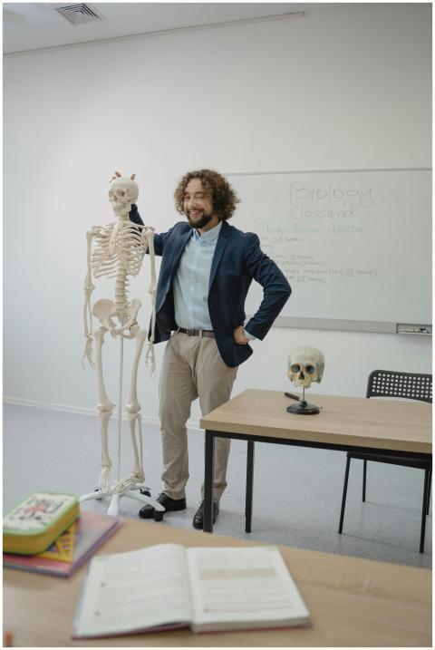 A smiling teacher stands beside a skeleton model i