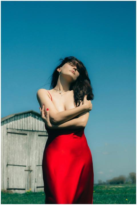 A woman in a red dress embraces herself outdoors,