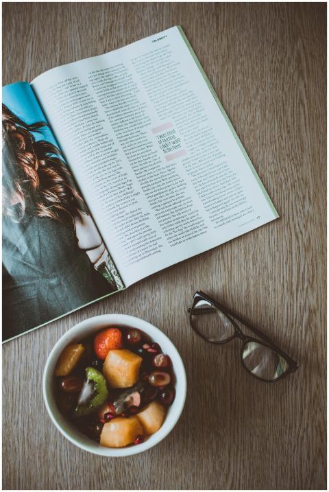A fresh fruit bowl, open magazine, and glasses on