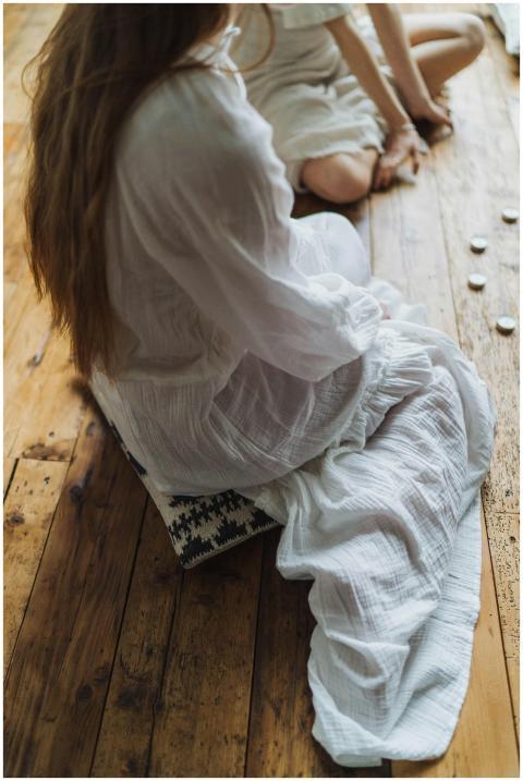 Two women in white sit on wooden floor, engaged in