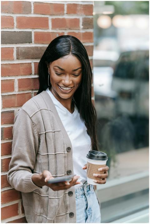Smiling woman stands by a brick wall, sipping coff