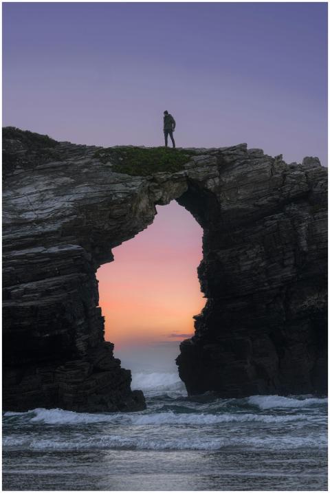 Man stands atop a stone arch overlooking the ocean