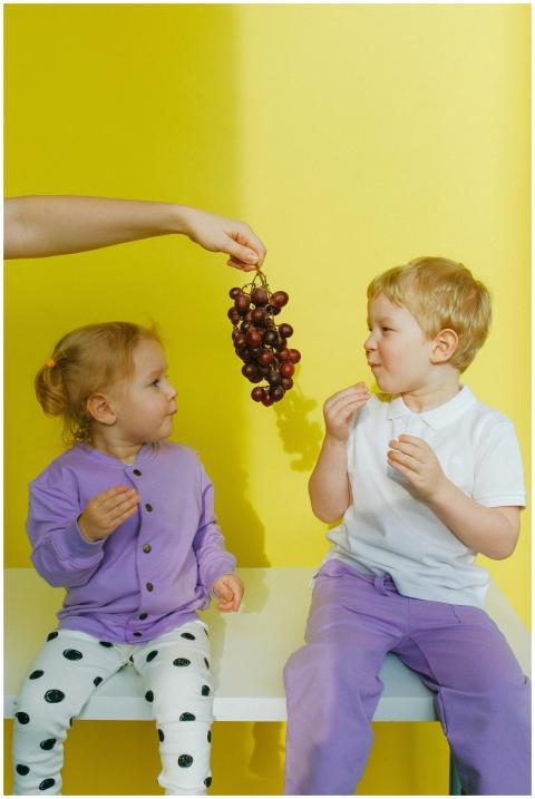 Two young children sitting indoors enjoying grapes
