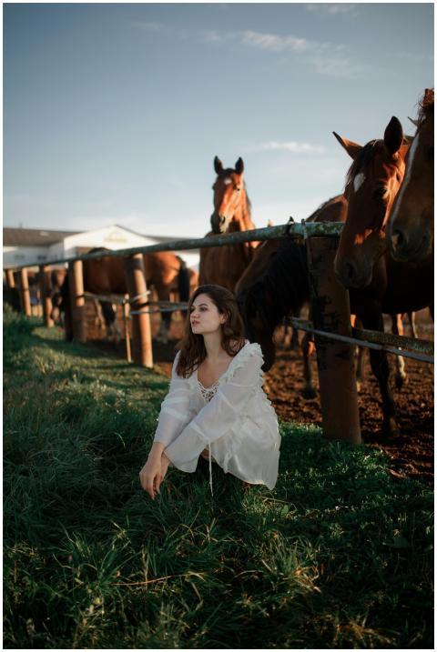 A woman in a white dress sits near a pen of horses