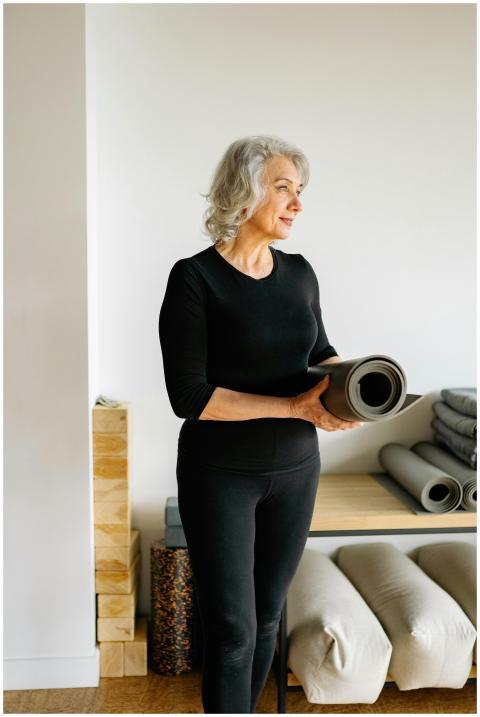 Elderly woman holding a yoga mat, standing in a br