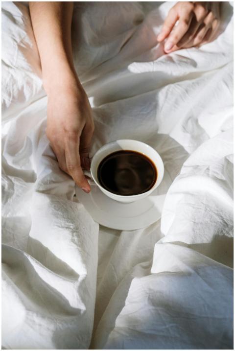 A person enjoys a cup of black coffee on white bed