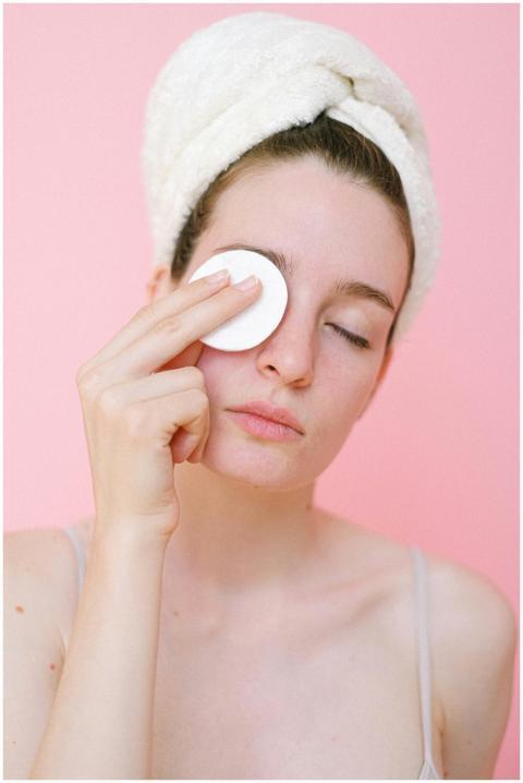 Young woman cleansing face with cotton pad on pink