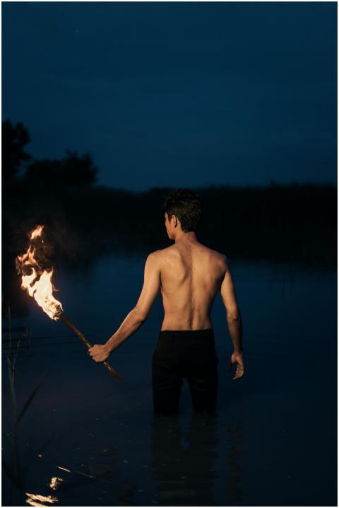 A topless man holds a fiery torch at night in calm