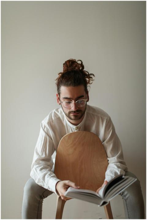 A young man with glasses sits on a chair reading a