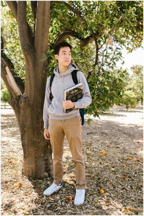 Asian teenage student standing by a tree holding b