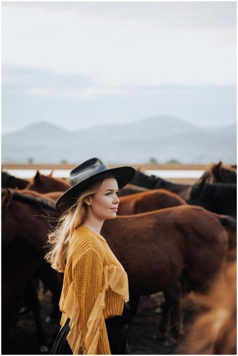 A woman in a stylish hat stands amidst a herd of h
