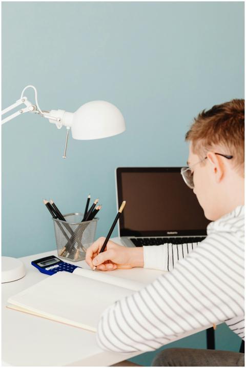Man writing in notebook at desk with laptop, lamp,